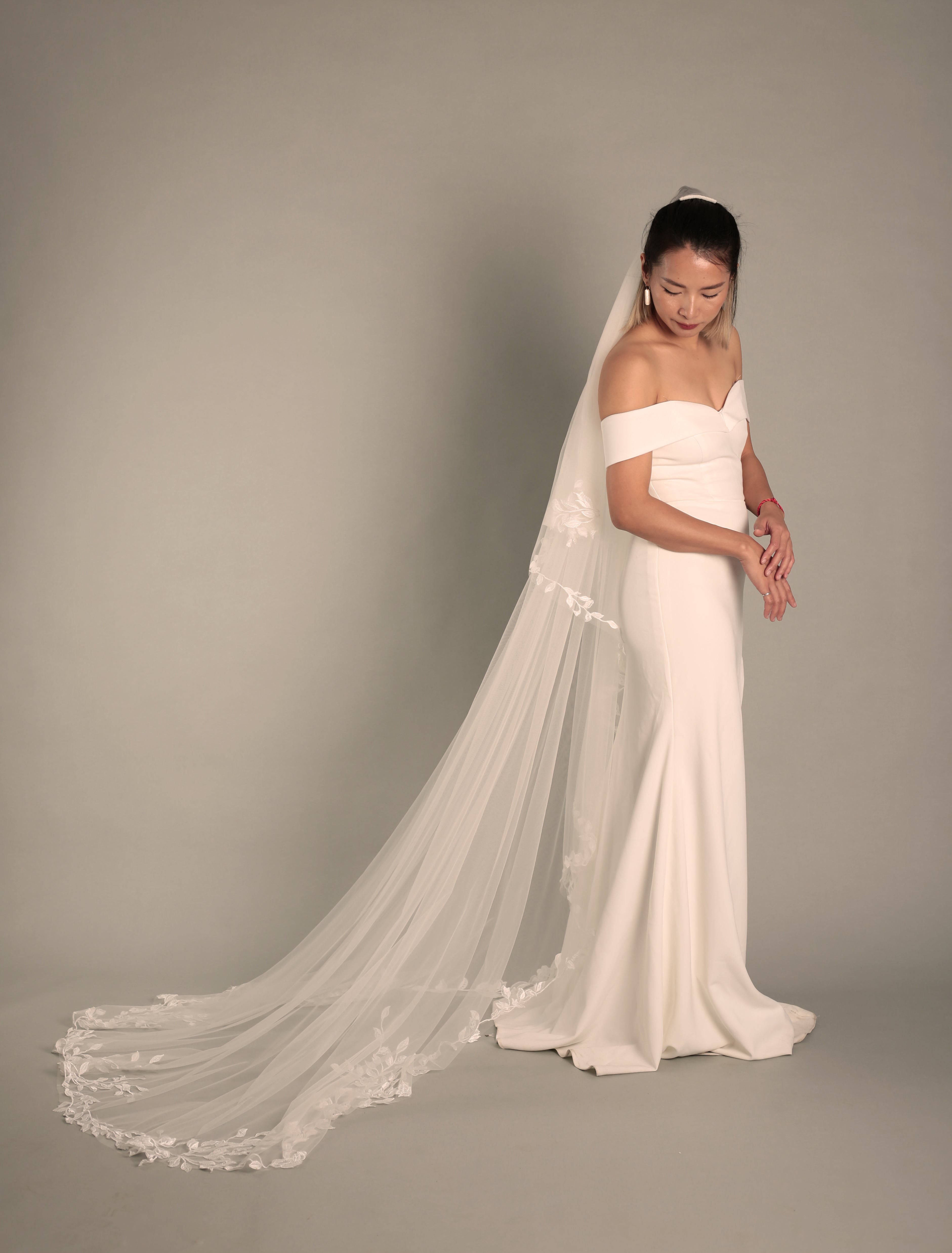 A bride wearing the BRITTNIE veil, captured from the right down side, showing the delicate floral lace along the edge and the graceful flow of the cathedral length.
