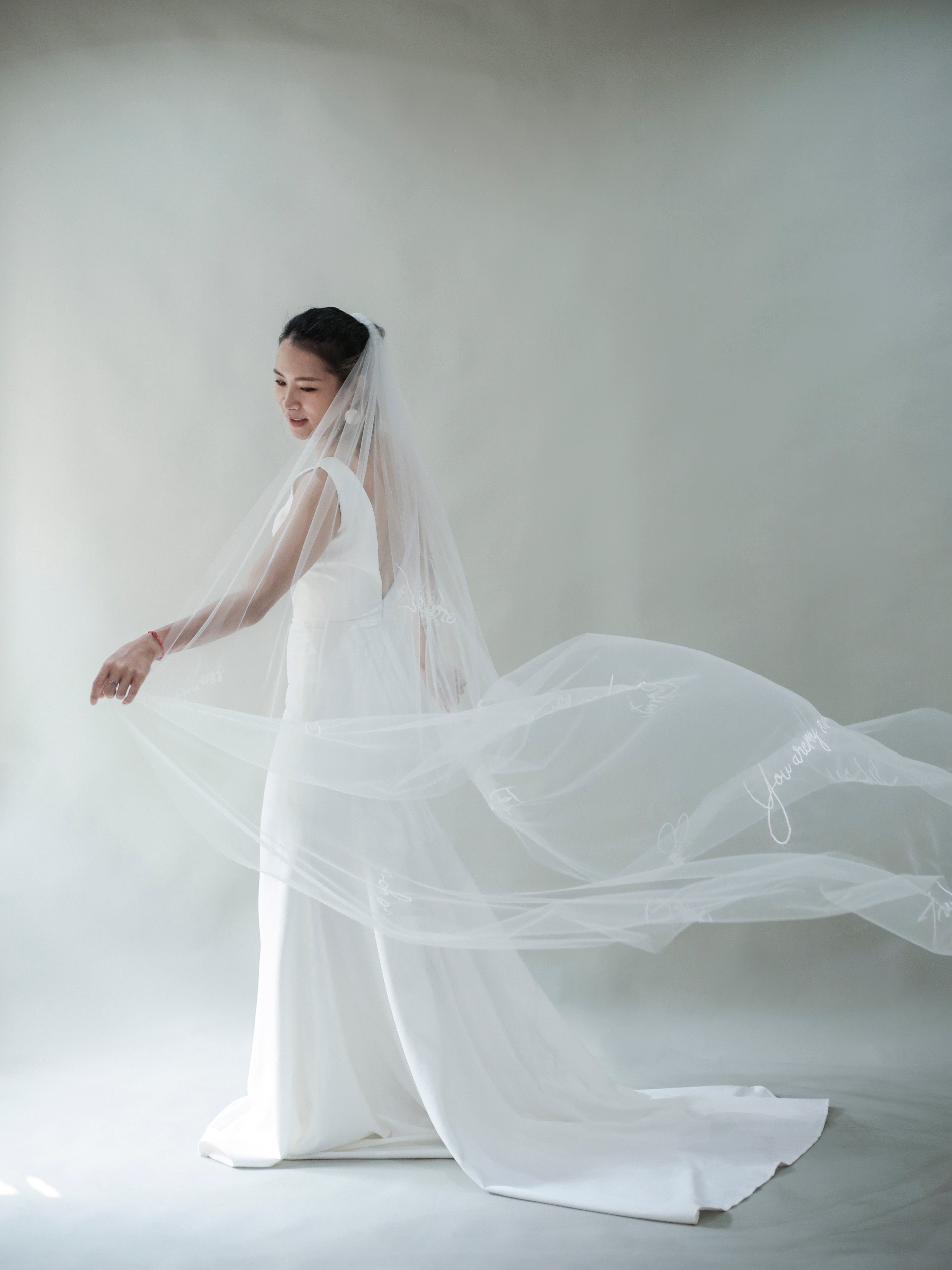 A bride wearing a white embroidered veil with light ivory color, standing in a chapel-like setting against a light background.