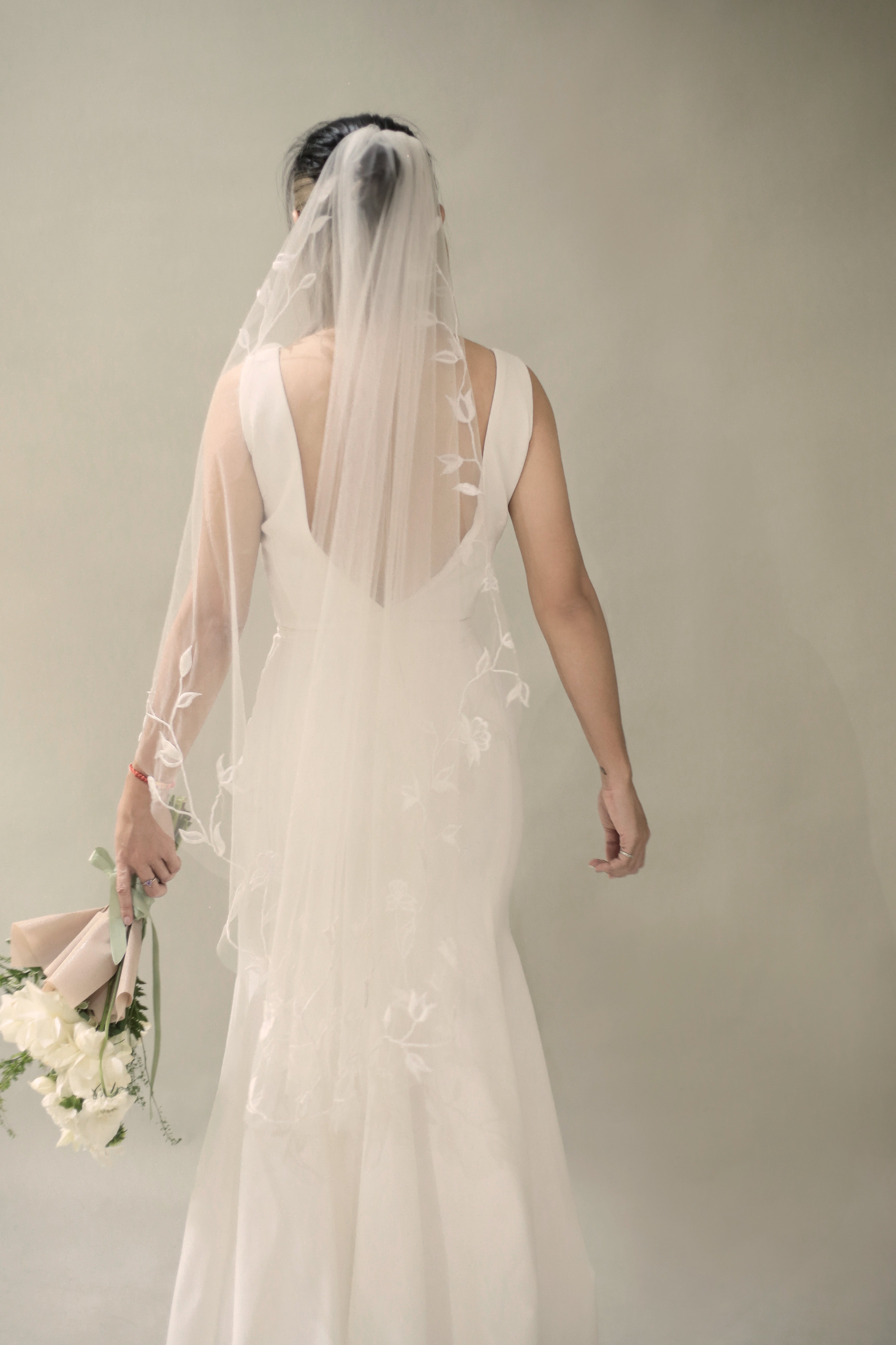 A bride wearing a white fingertip veil with floral embroidery, walking away from the camera holding a bouquet.