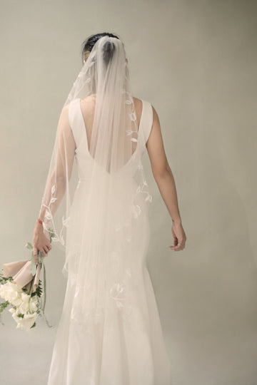 A bride wearing a white fingertip veil with floral embroidery, walking away from the camera holding a bouquet.