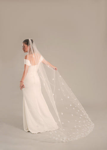 A woman wearing a white bridal gown and a petal chapel veil that extends into a long train, standing in a graceful pose.
