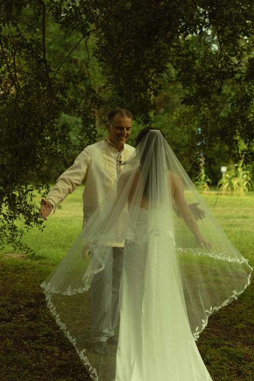 Bride Mckhayla wearing a cathedral-length lace floral veil in a vintage bohemian garden theme wedding kissing under an old giant tree