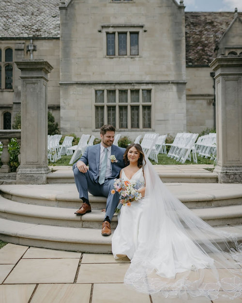 Bride in white gown and groom in blue suit sitting on stone steps of a historic building before their wedding ceremony featuring cathedral floral wedding veil