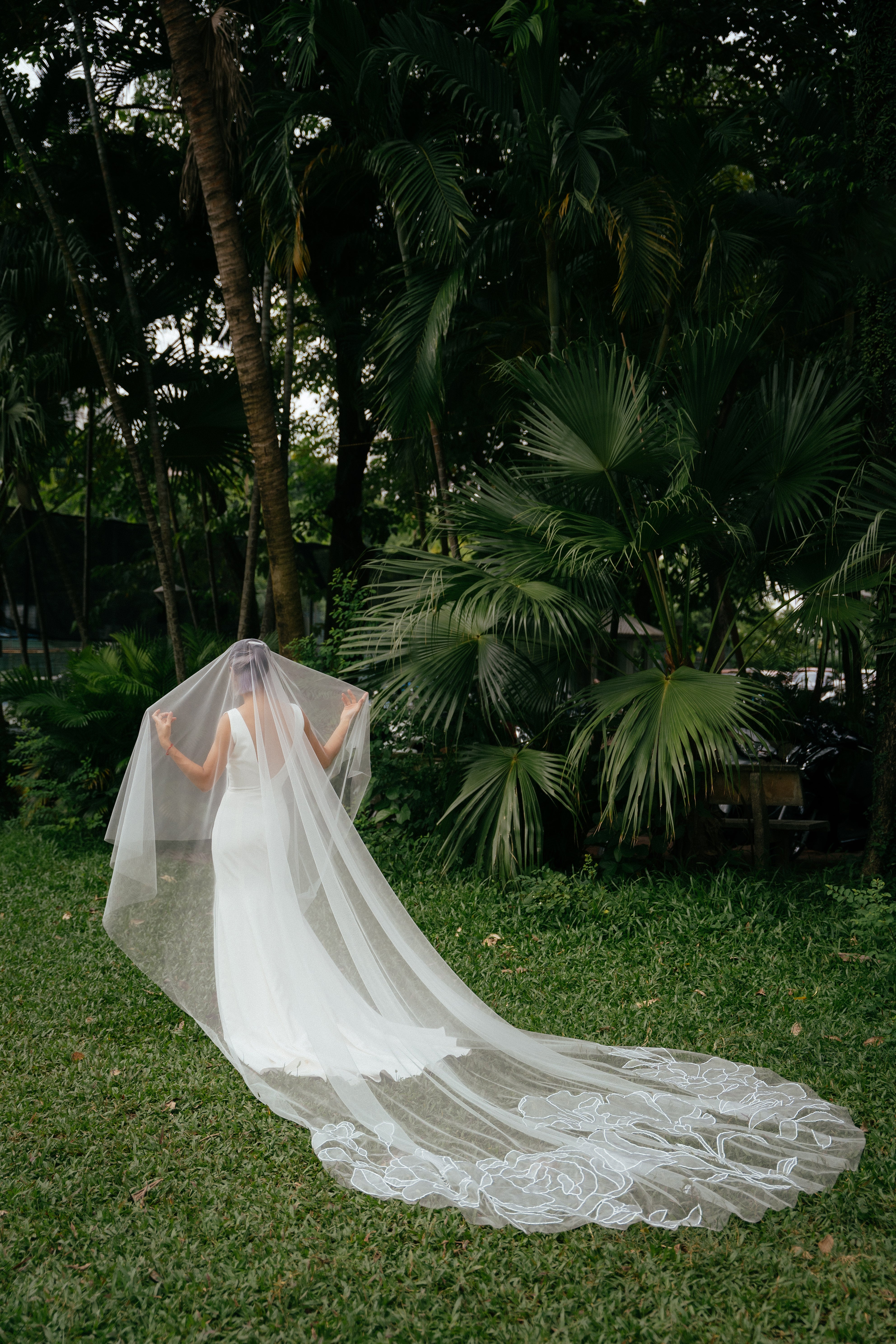 A bride in a grassy outdoor setting wearing a white cathedral-length wedding veil with floral embroidery, standing near palm trees.