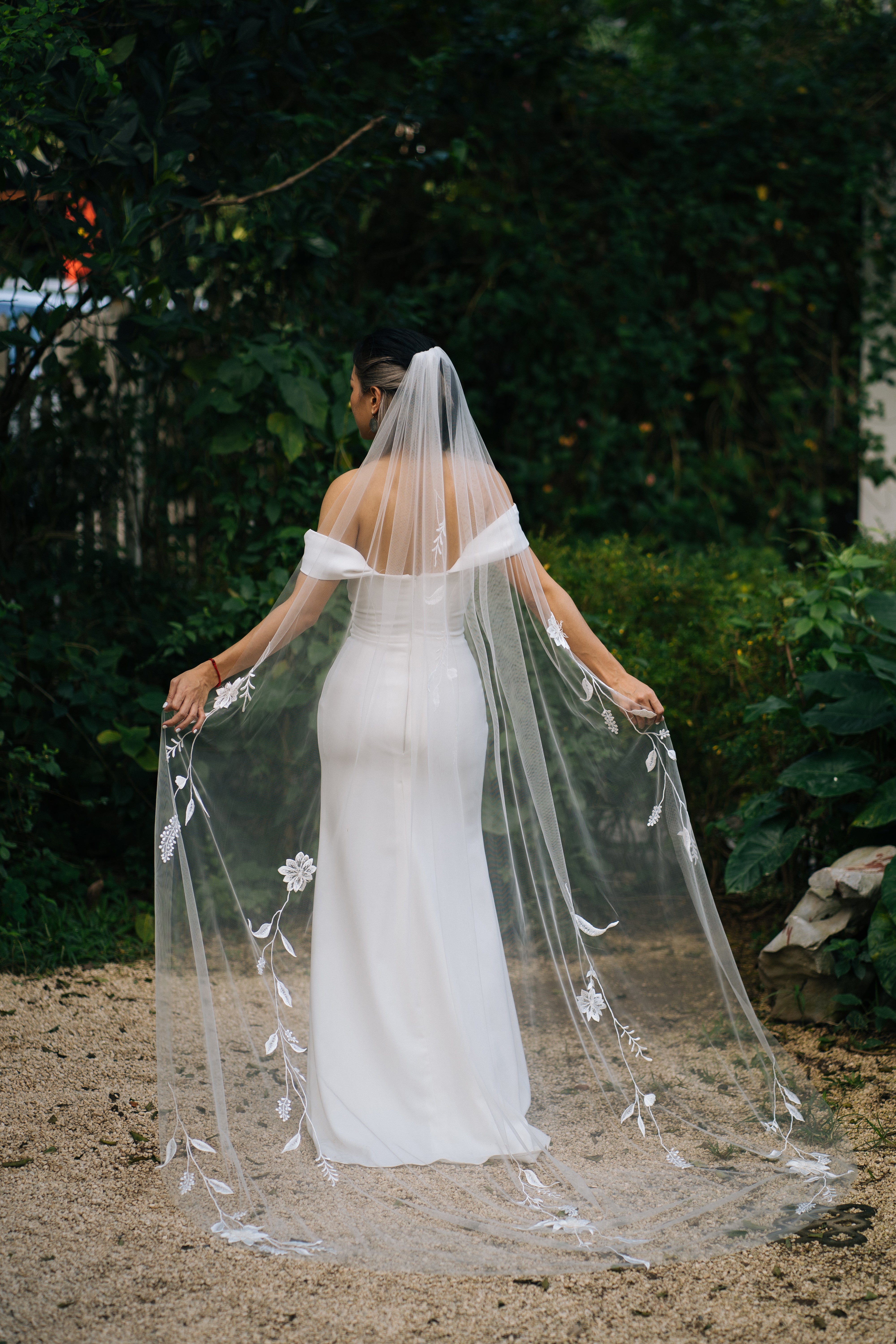 A bride wearing a white embroidered wedding veil with 3D flowers, standing outdoors with trees and greenery in the background.