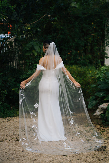 A bride wearing a white embroidered wedding veil with 3D flowers, standing outdoors with trees and greenery in the background.