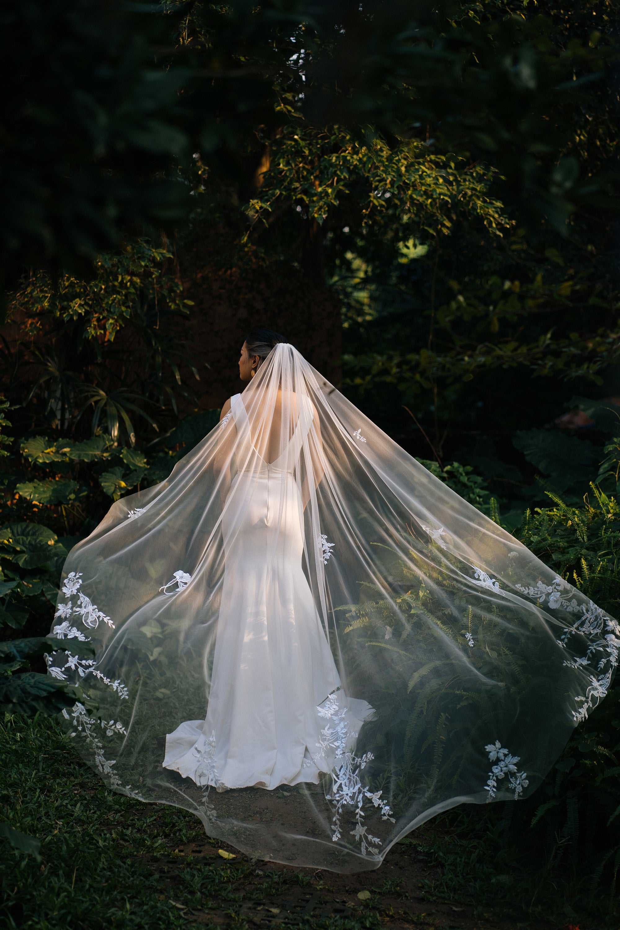 A bride wearing a white floral lace wedding veil that extends to the ground, standing in a garden setting.