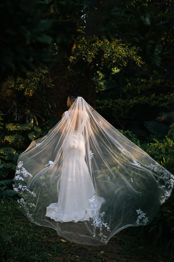 A bride wearing a white floral lace wedding veil that extends to the ground, standing in a garden setting.