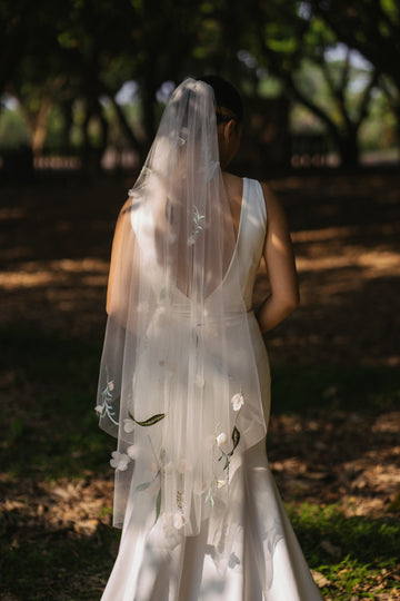 A bride wearing a white wedding veil with 3D floral embroidery in soft colors standing in an outdoor setting.