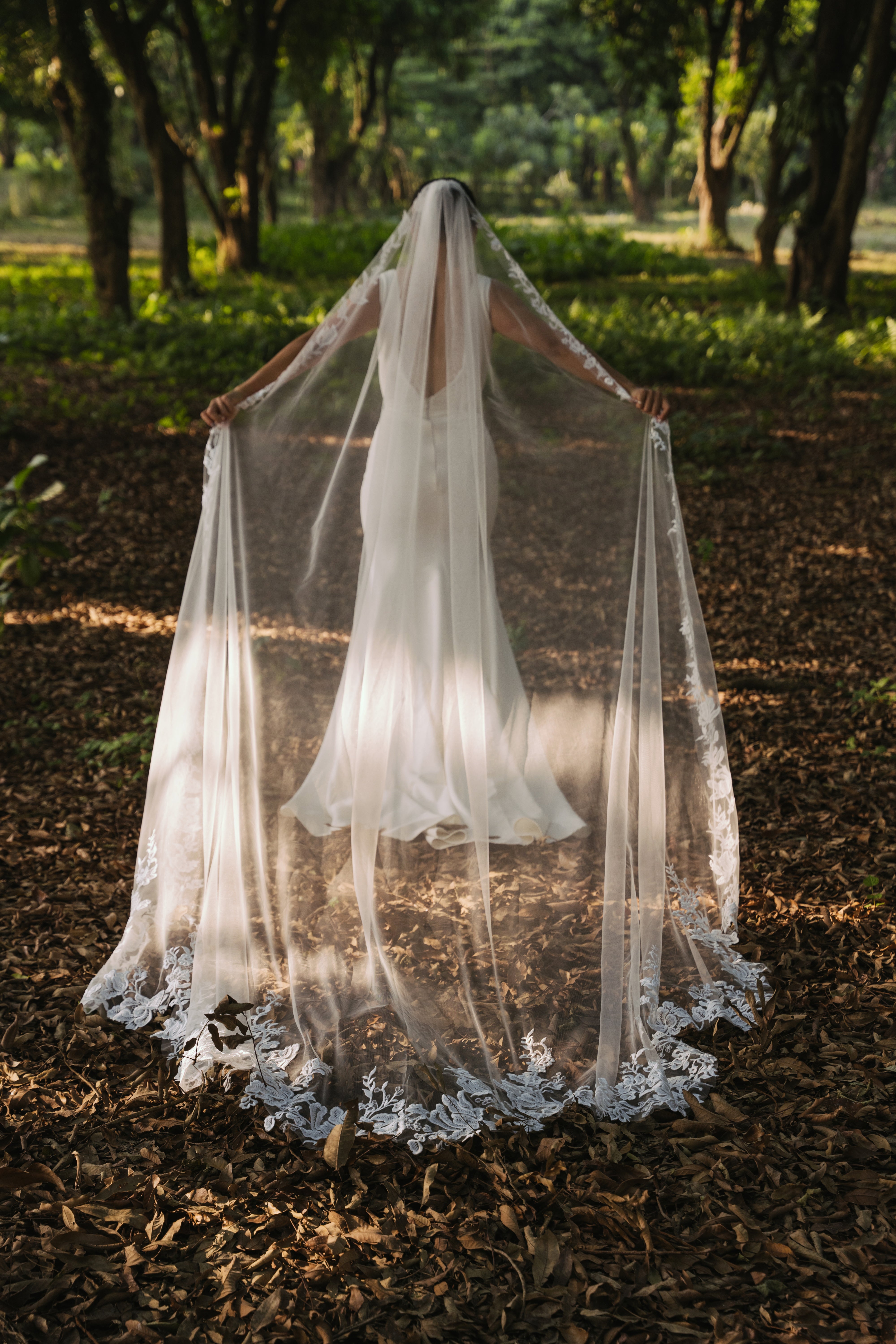 A bride wearing a white cathedral-length lace wedding veil with delicate lace roses and a unique flower-like shape at the train, standing in a forest setting.