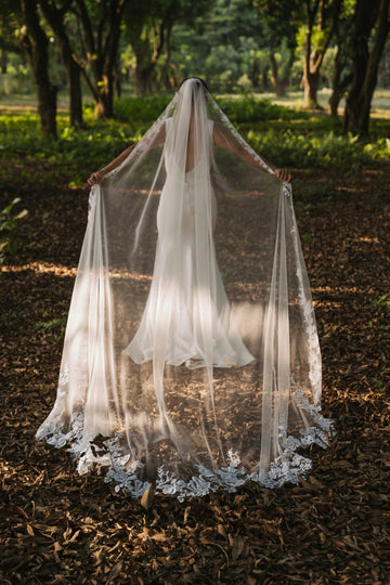 A bride wearing a white cathedral-length lace wedding veil with delicate lace roses and a unique flower-like shape at the train, standing in a forest setting.
