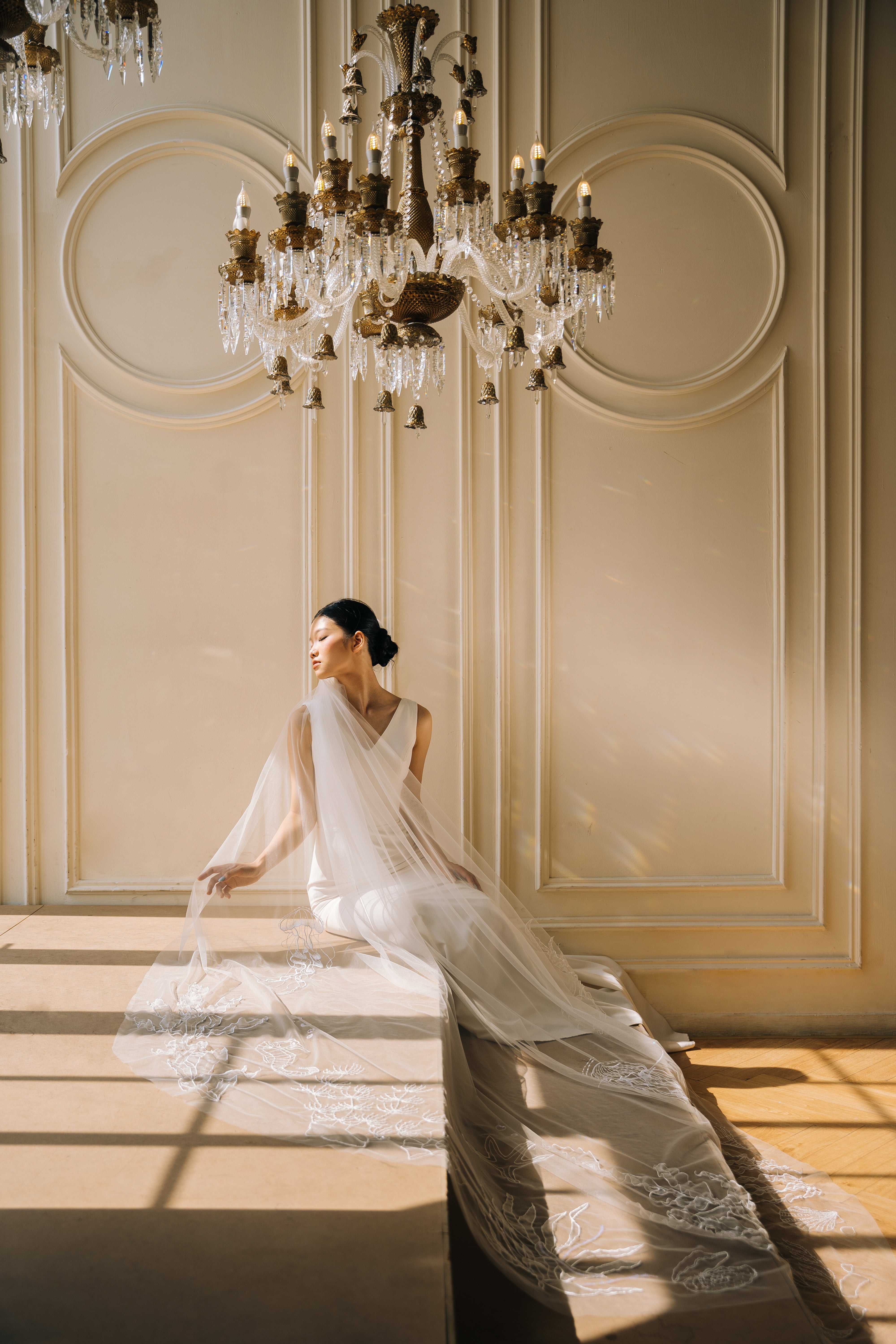 A model wearing a light ivory, cathedral-length wedding veil with 3D floral embroidery, standing in a room with a chandelier.