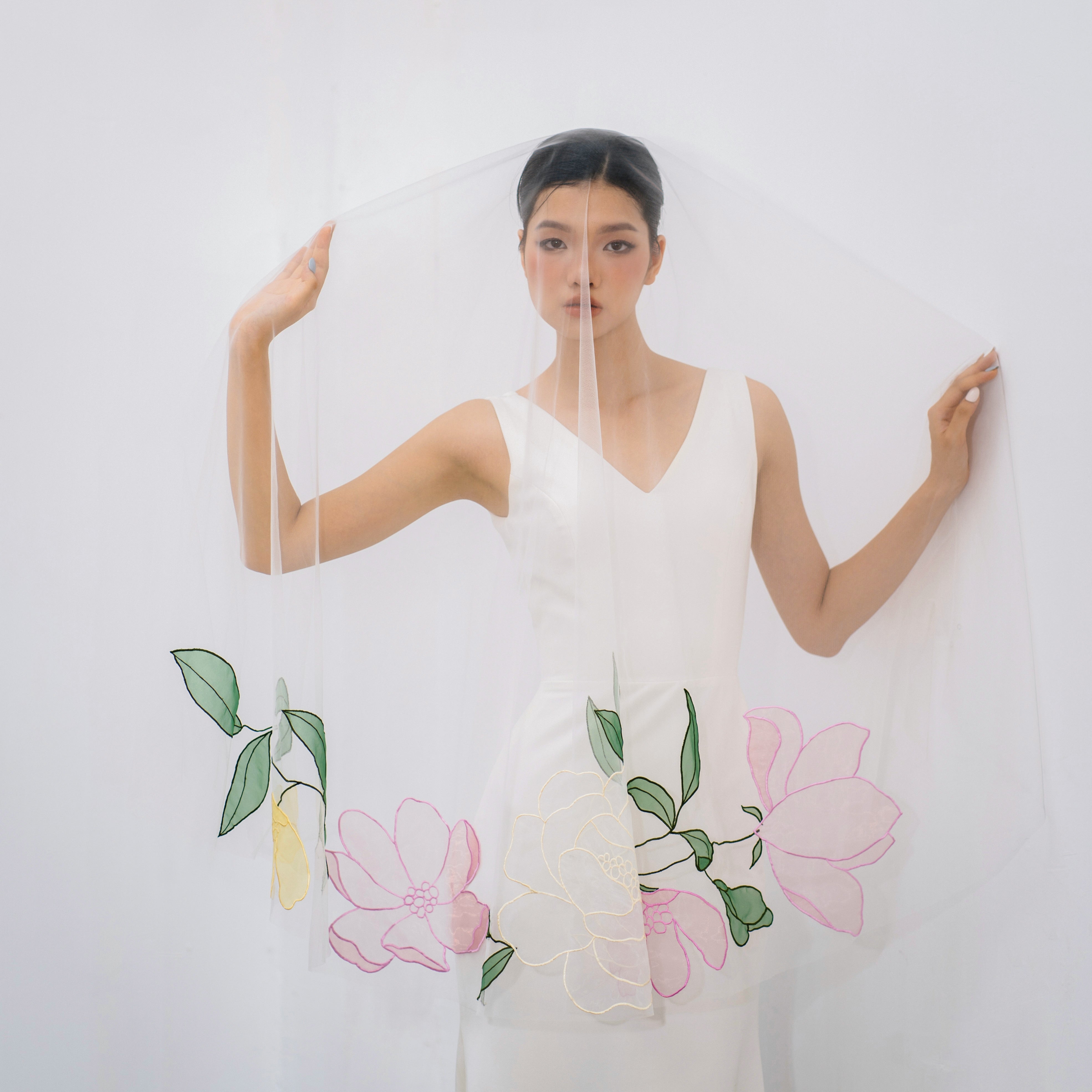 A woman wearing a white fingertip veil with embroidery floral designs in pink, pale yellow, and green, standing in front of a white background.