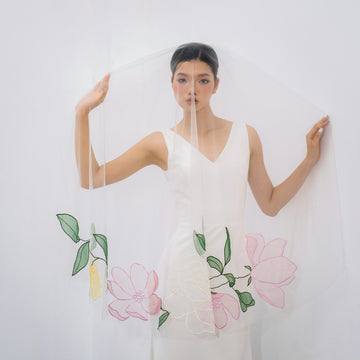 A woman wearing a white fingertip veil with embroidery floral designs in pink, pale yellow, and green, standing in front of a white background.