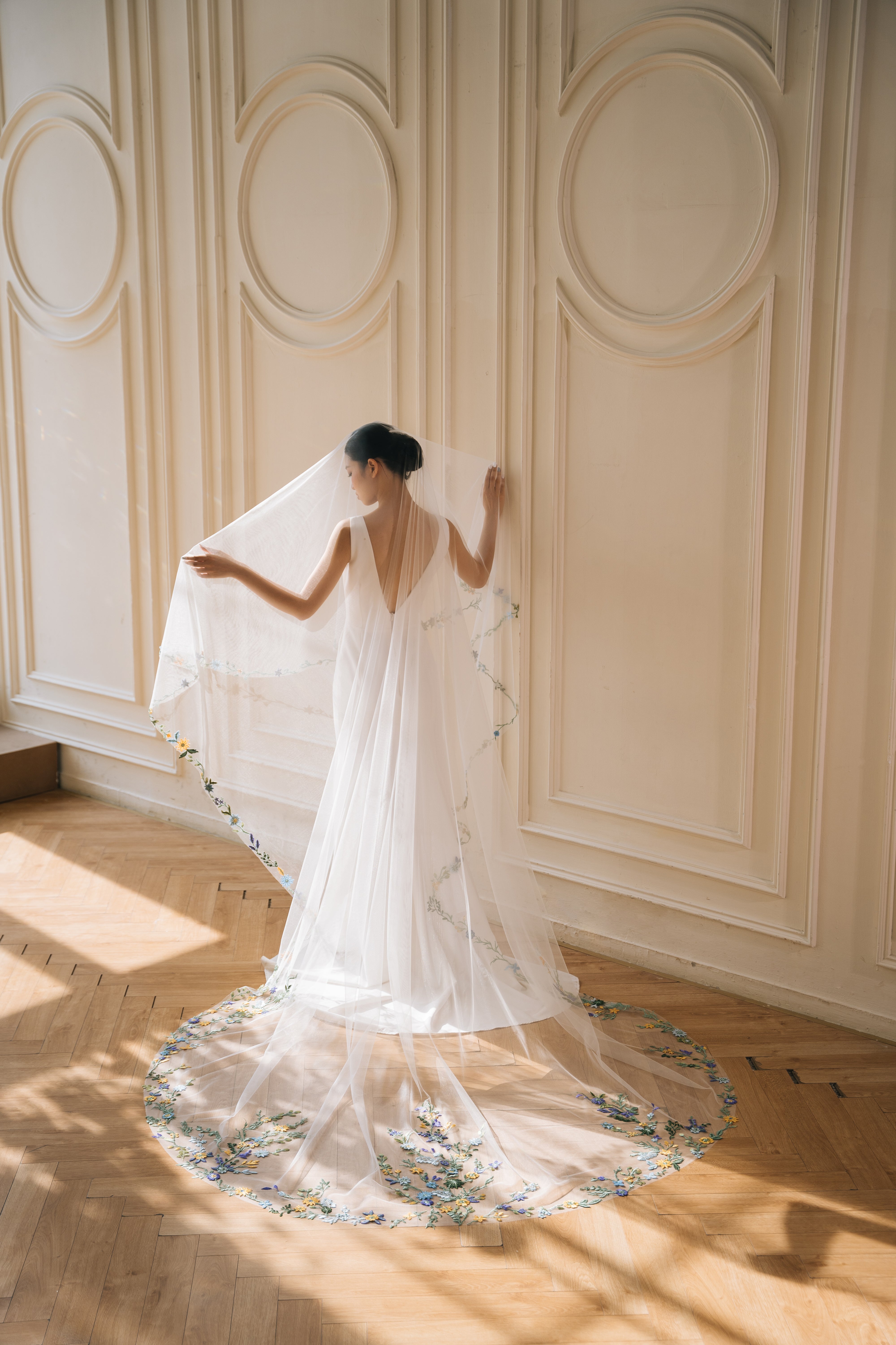 A bride in a white wedding veil with intricate floral lace detailing, standing in a room with elegant decor.
