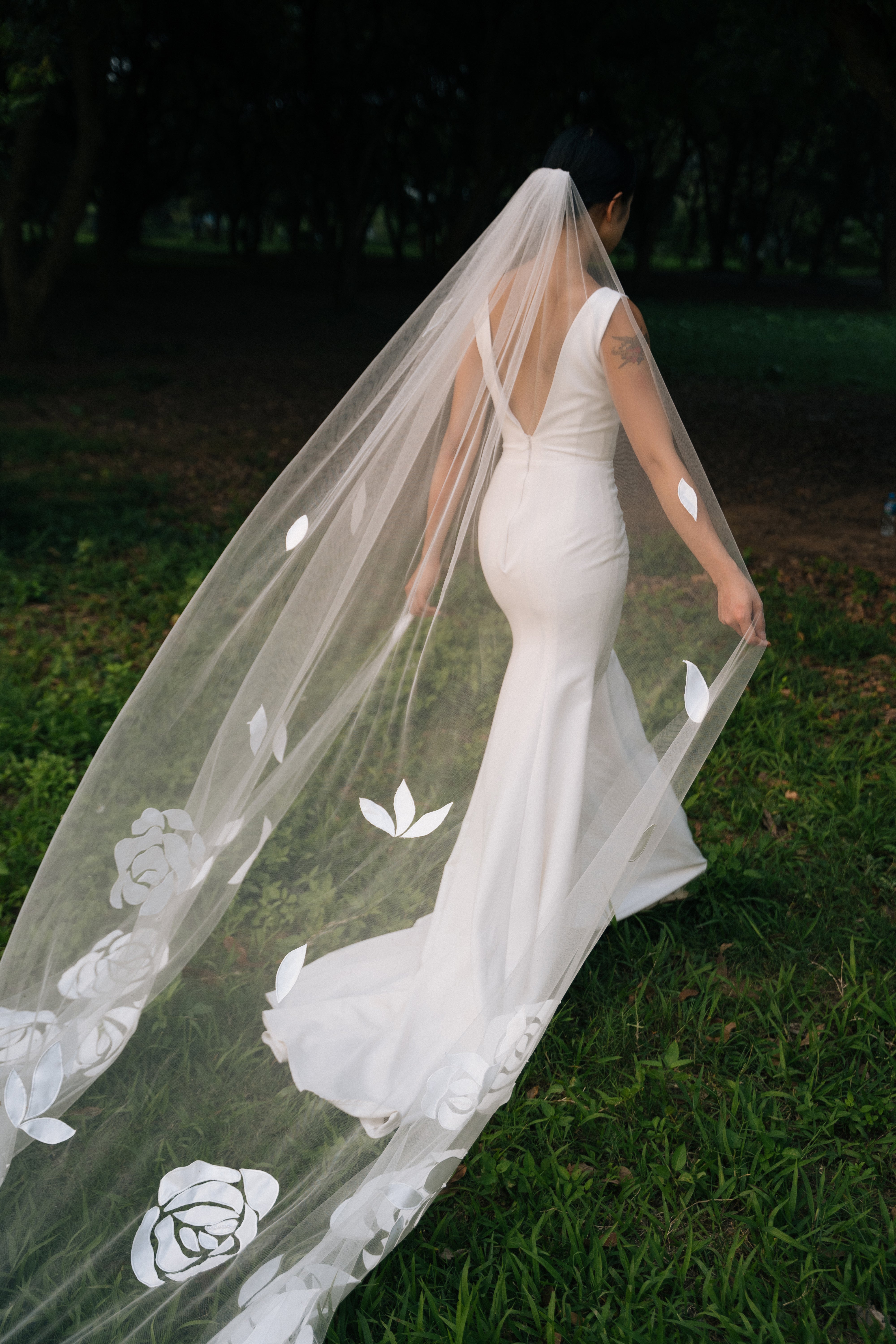 A bride wearing a white cathedral-length wedding veil with embroidered roses and leaves, walking outdoors.
