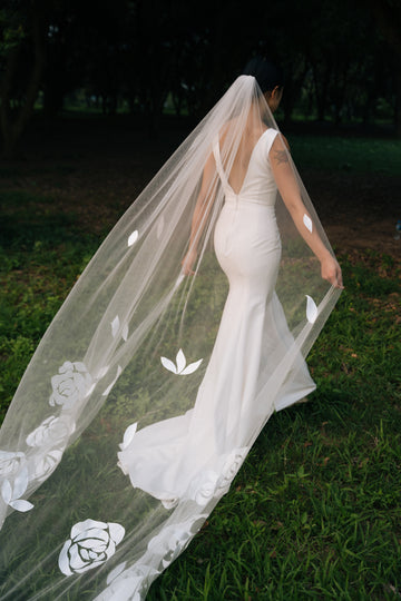 A bride wearing a white cathedral-length wedding veil with embroidered roses and leaves, walking outdoors.
