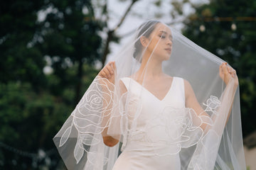 A bride holding a cathedral-length wedding veil with floral embroidery, standing outdoors.