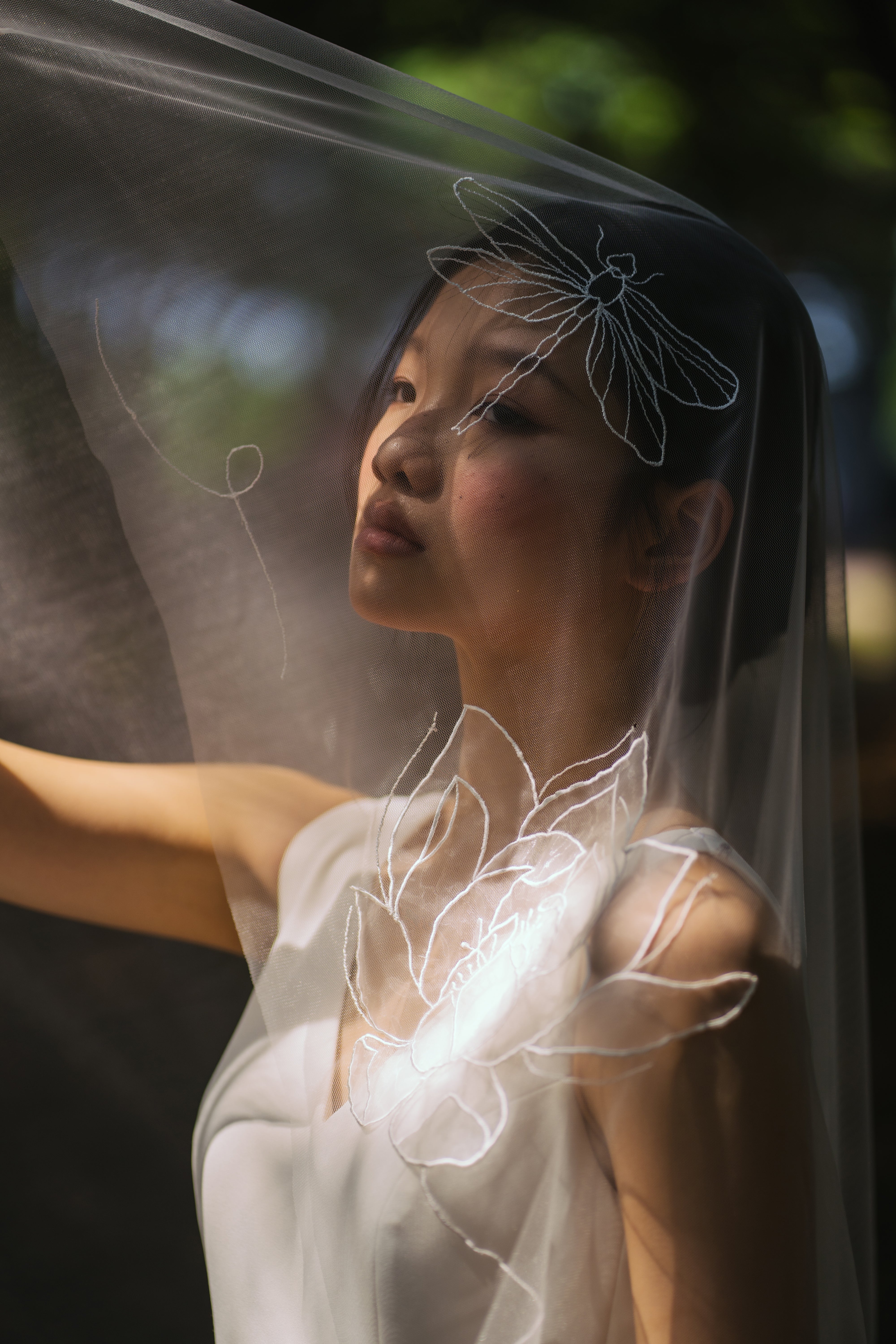 A woman wearing a white bridal veil with embroidered floral patterns and dragonfly motifs, standing outdoors with a blurred background.