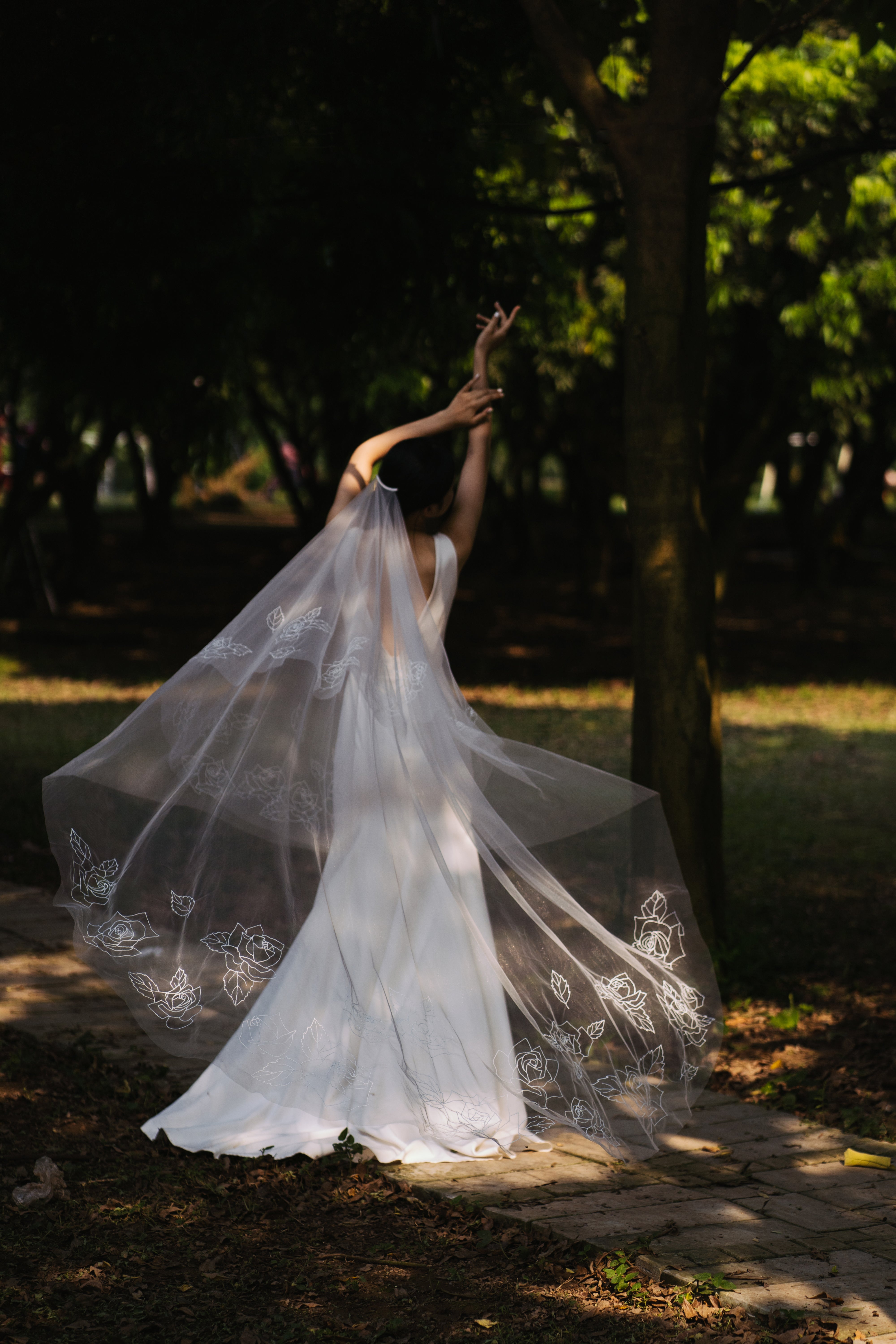 A woman wearing a light ivory, floral embroidered bridal veil that extends into a chapel length, swirling in a park with trees in the background.