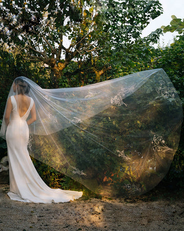 A bride wearing a white chapel-length wedding veil with embroidered wildflower designs, standing outdoors with greenery in the background.