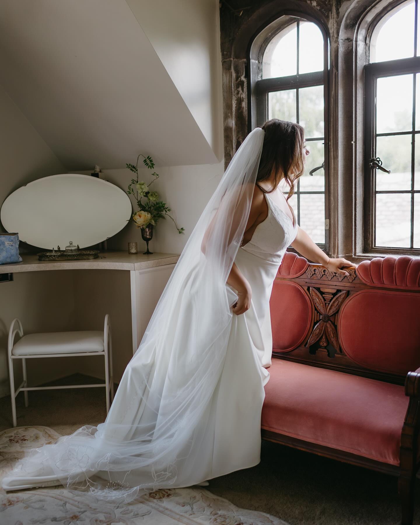 Woman in a white dress with a long veil in a room with a red couch and large windows.
