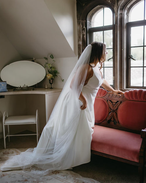 Woman in a white dress with a long veil in a room with a red couch and large windows.