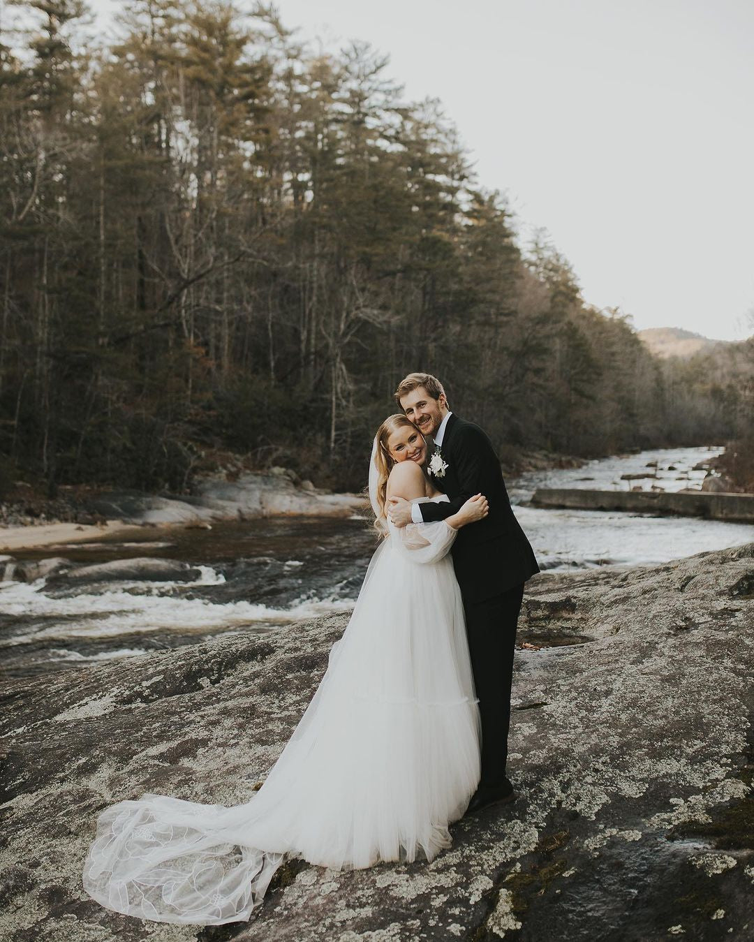 A bride and groom standing on a rock in front of a river